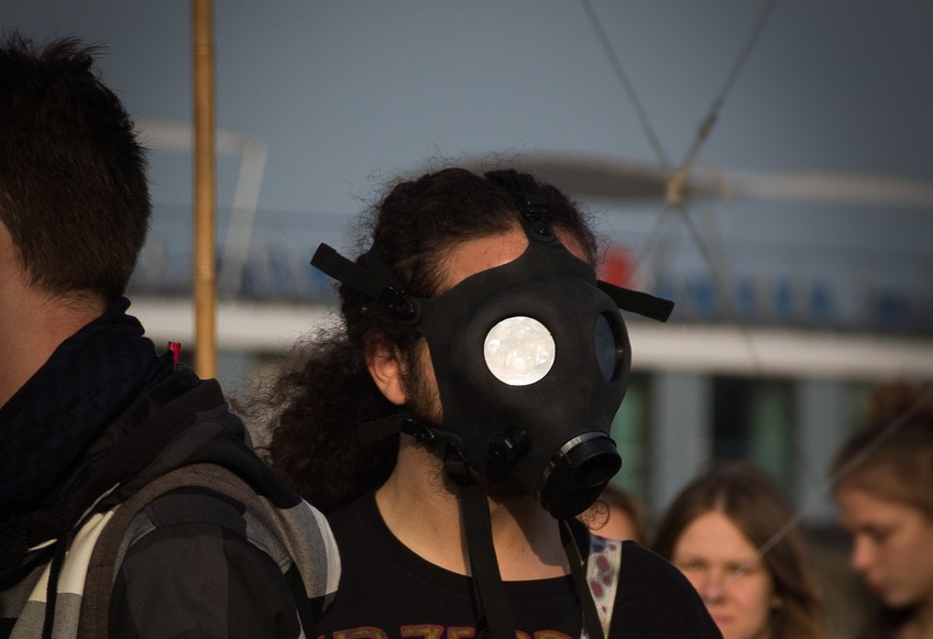 A white-figured person with long curly dark hair is at the centre of the image with their face obscured by a gas mask. There are four other white-figured people visible but obscured in the image. They aer outside in daylight but the background is also obscured