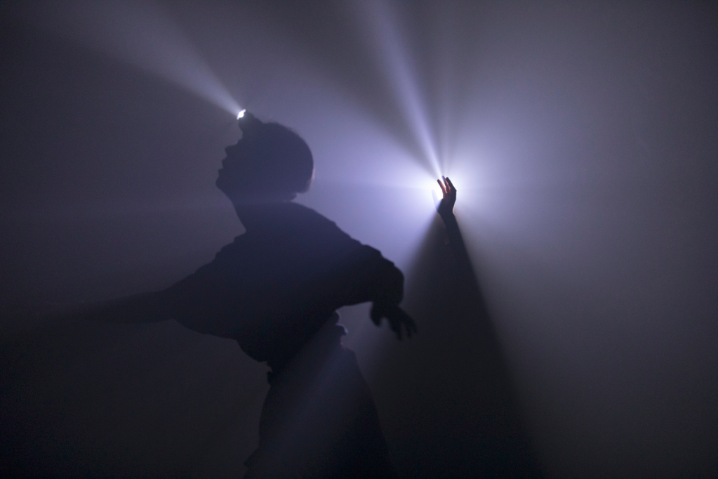 A shadowy figure wearing a head torch is silhouetted against the darkness, facing towards the left, their left arm outstretched towards the edge of the frame, their right arm stretched towards the centre. Behind them a hand emerges from a triancle of darkness, light dispersing from their fingertips